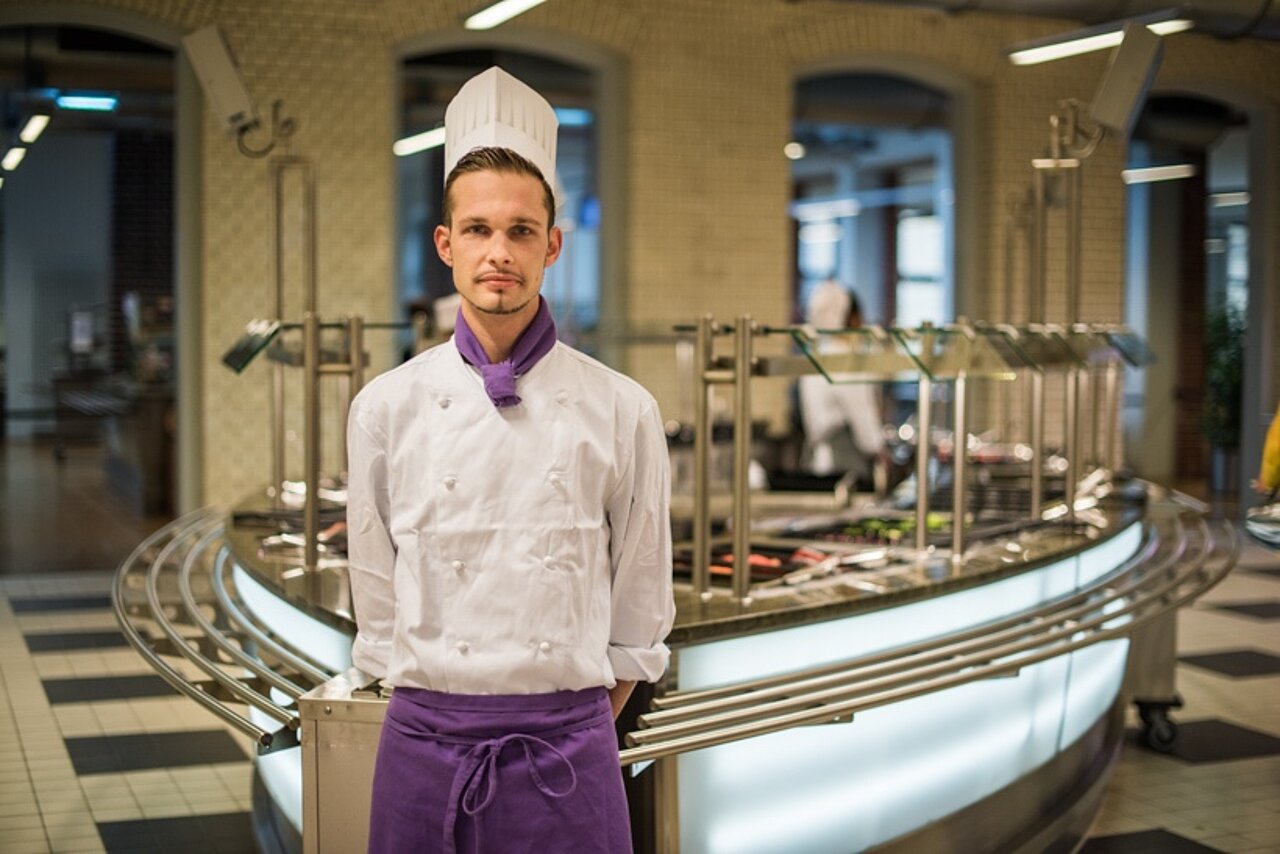 Auszubildender in weißer Kochjacke und Kochmütze steht vor der Essensausgabe in einer Mensa.