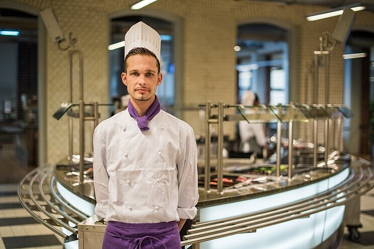 Auszubildender in weißer Kochjacke und Kochmütze steht vor der Essensausgabe in einer Mensa.