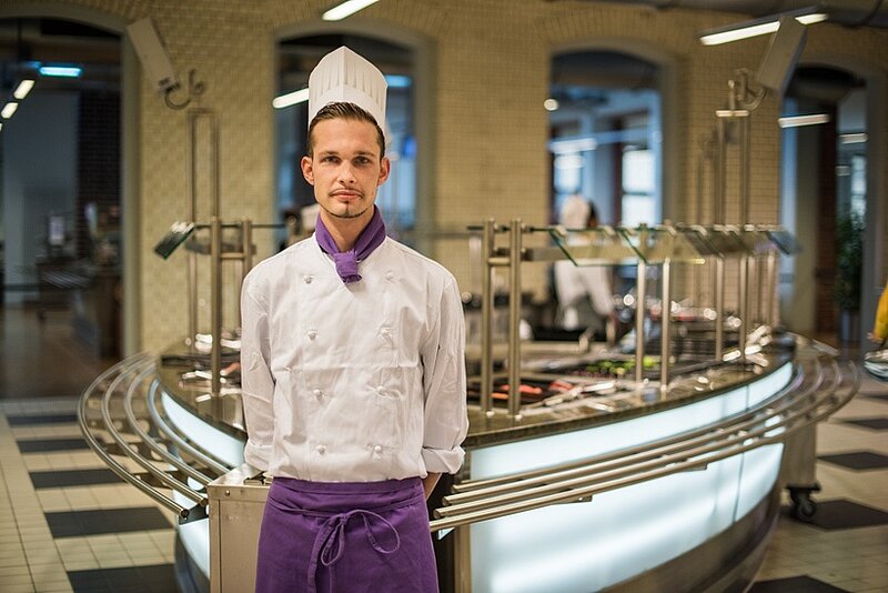 Auszubildender in weißer Kochjacke und Kochmütze steht vor der Essensausgabe in einer Mensa.