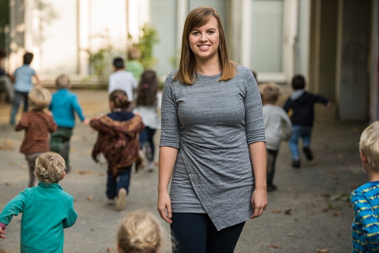 Erzieherin steht vor einer Kita, während Kinder im Hintergrund spielen.