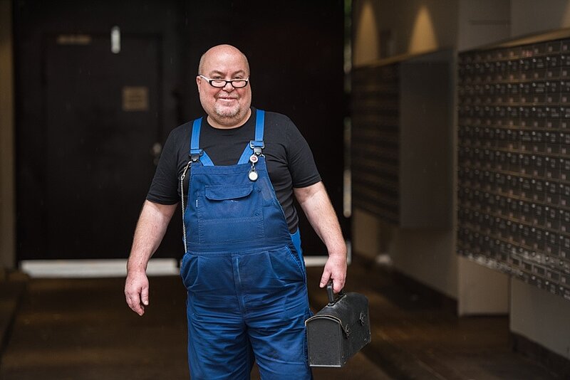 Hausmeister in blauer Arbeitslatzhose vor einem Wohnheim.